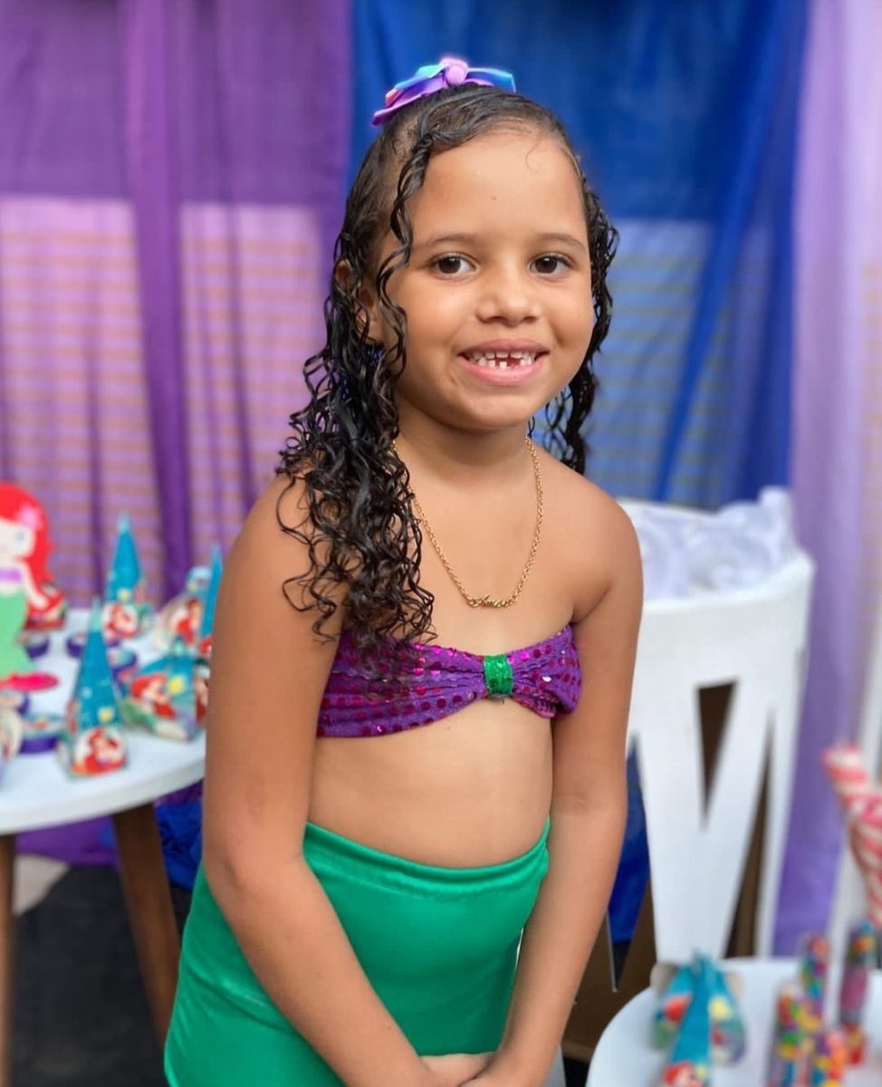 Heloysa era uma criança negra de cabelos castanhos e cacheados; na foto ela está vestindo um top roxo e uma saia verde, combinando com a decoração de festa de aniversário ao fundo; ela está sorrindo