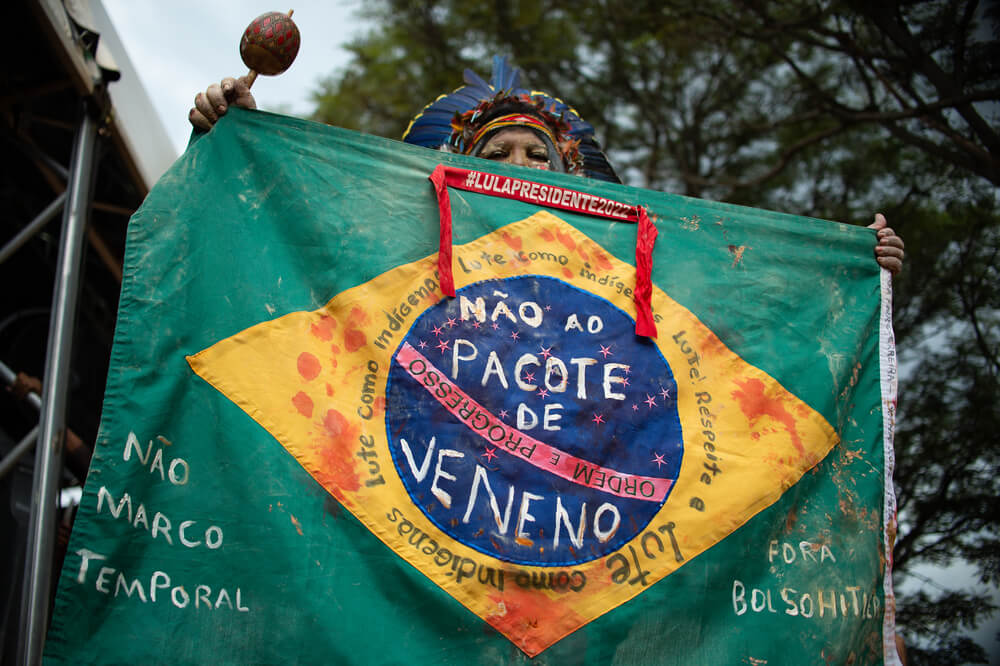 Indígena segura a bandeira do Brasil nas mãos; ela está manchada de sangue e com algumas frases escritas como "não ao pacote do veneno", "Fora Bolsonaro" e "Não ao Marco Temporal"