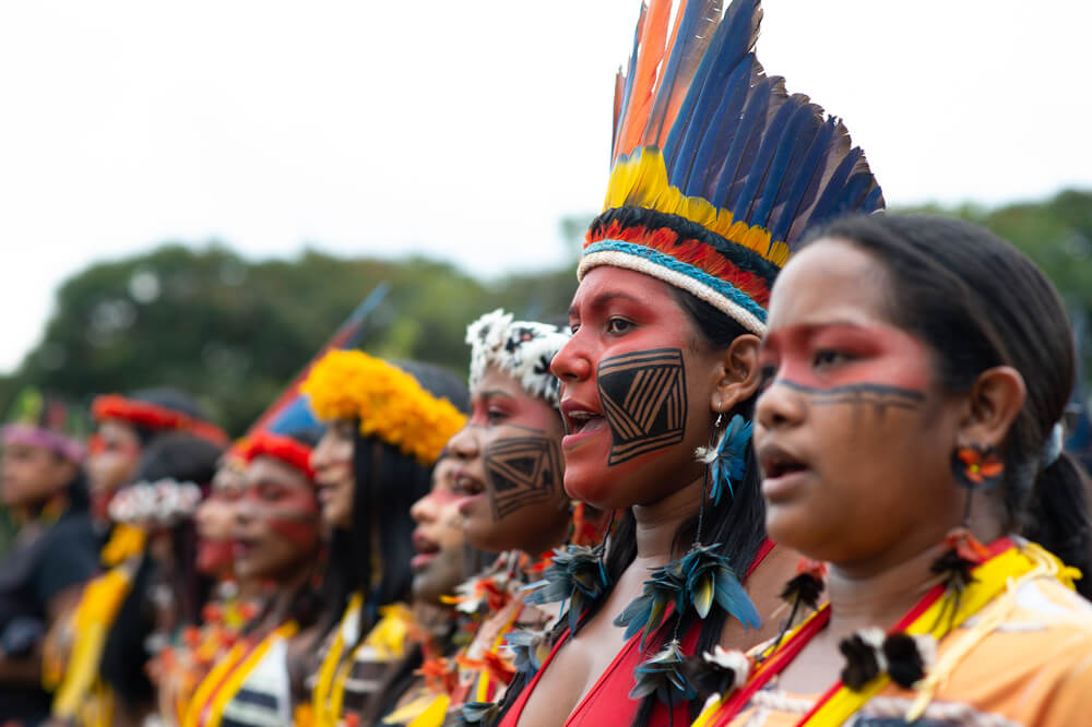 Jovens mulheres indígenas organizadas em fileira durante ato do ATL 2022. Elas vestem cocar e outros adereços tradicionais, além de pintura facial étnica