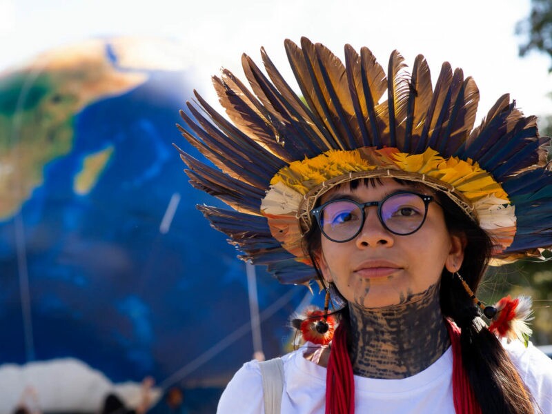 Txai é uma jovem indígena com cabelos e olhos castanhos. Ela usa óculos, um cocar na cabeça, brinco de pena nas orelhas e tem pintura étnica no rosto e pescoço. Atrás dela há um balão azul imitando a forma do planeta terra.