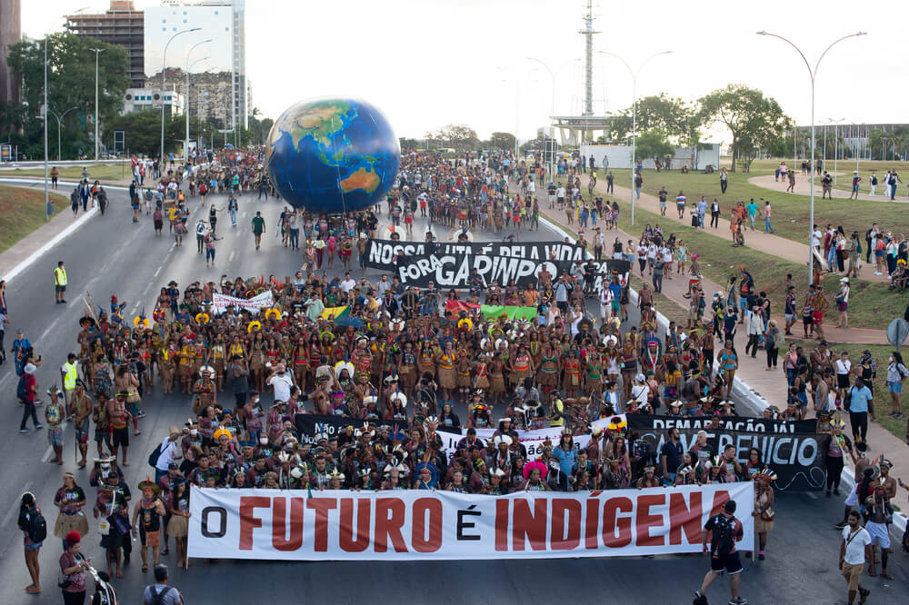 Imagem mostra dezenas de indígenas durante passeata do ATL 2022. É possível ver algumas faixas e cartazes que eles seguram, como a em destaque na frente do grupo com a frase 