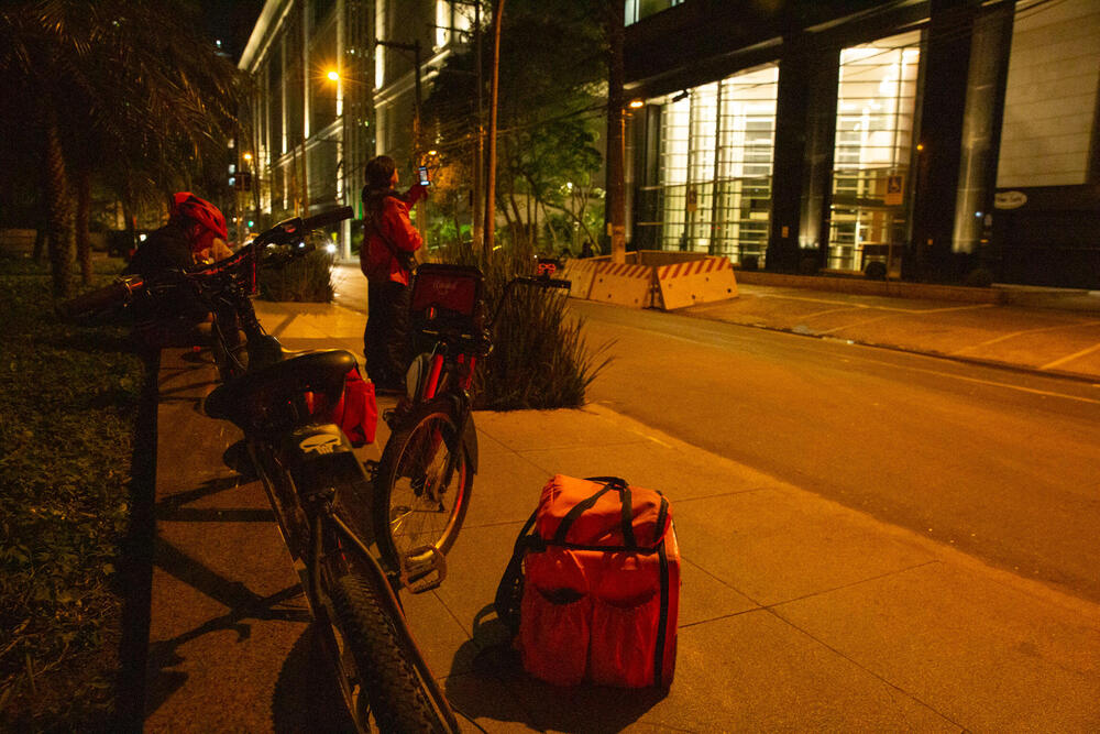 Imagem noturna mostra bags de entregadores do iFood paradas em uma calçada em frente a um prédio comercial