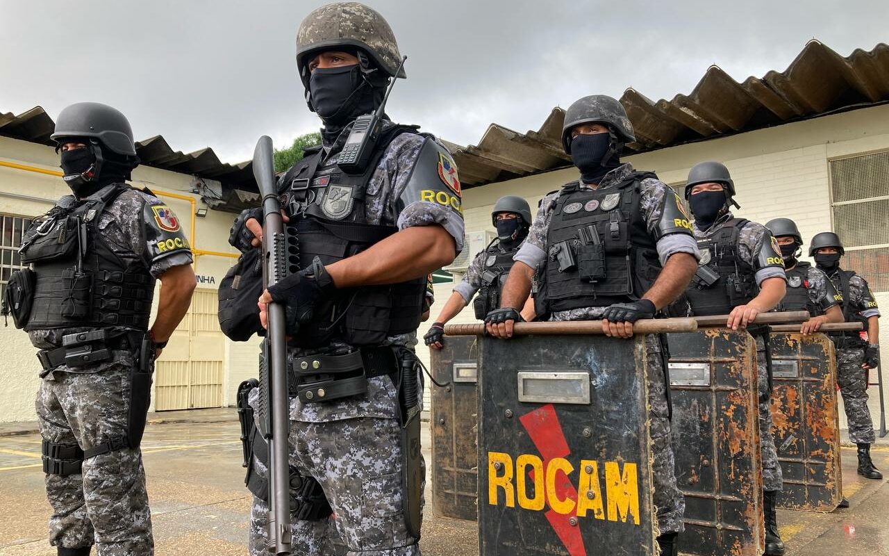 Policiais da ROCAM, trajados com uniforme, colete, capacete e armas, em frente à presídio no Amazonas
