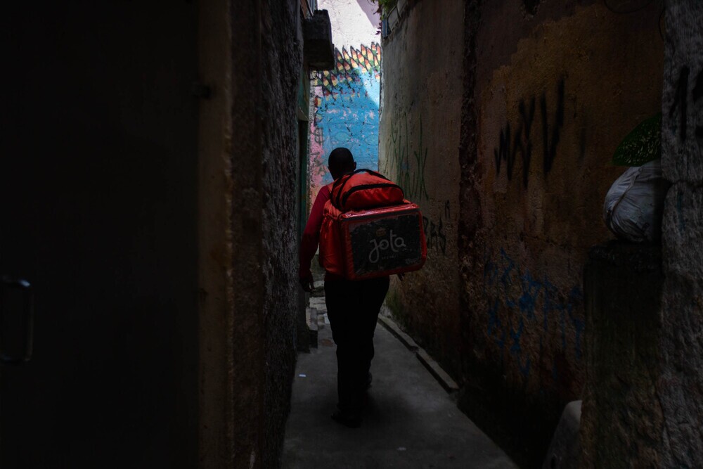 Entregador com bag do iFood nas costas caminha entre beco