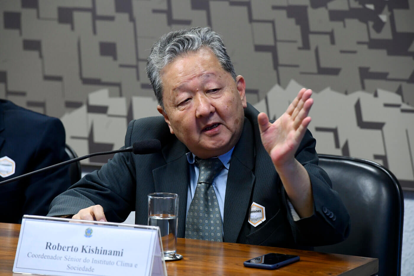 Roberto Kishinami é um homem asiático, com cabelos grisalhos e rosto redondo; Roberto usa um terno preto com camisa social azul e gravata cinza