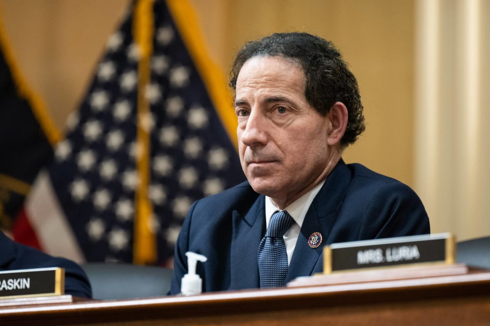 O Congressista Jamie Raskin é um homem branco, com cabelos e olhos escuros. Raskin veste terno e gravata, e aparece em frente â bandeira dos EUA.