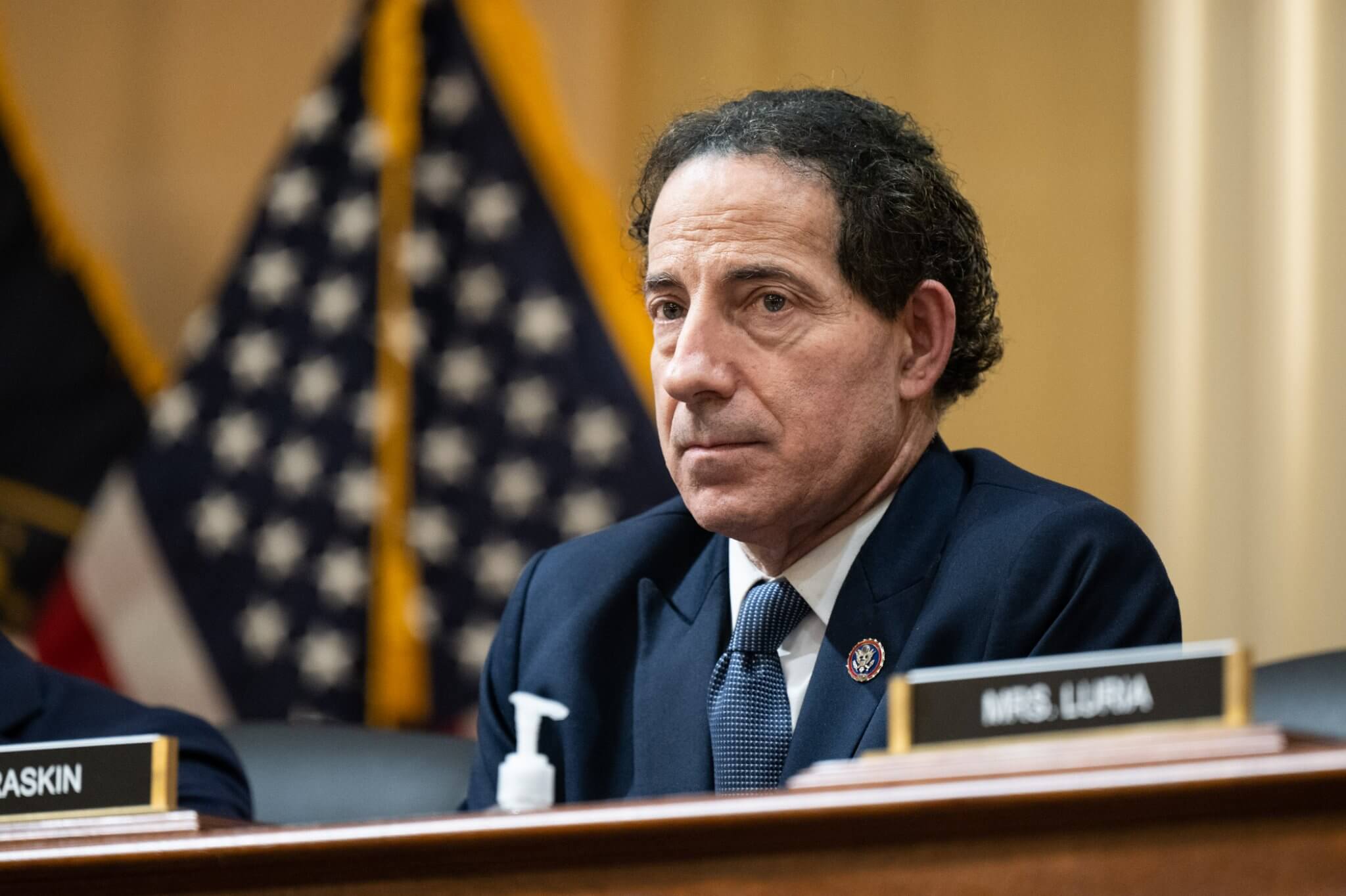 O Congressista Jamie Raskin é um homem branco, com cabelos e olhos escuros. Raskin veste terno e gravata, e aparece em frente â bandeira dos EUA.