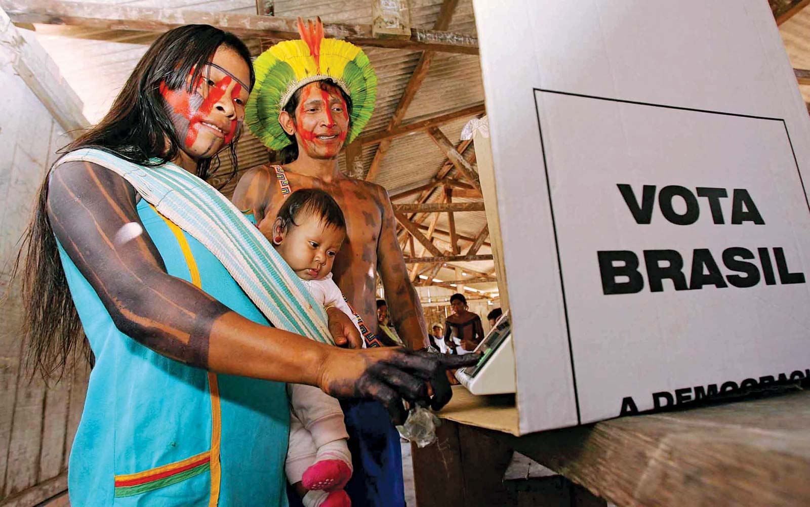 Casal indígena com bebê em aldeia em cabina de votação realiza seu voto.