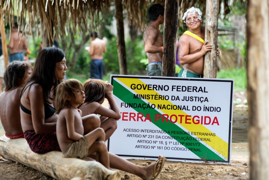 Indígenas yanomamis sentados ao lado de placa da Funai que atesta o território como terra protegida pelo governo federal