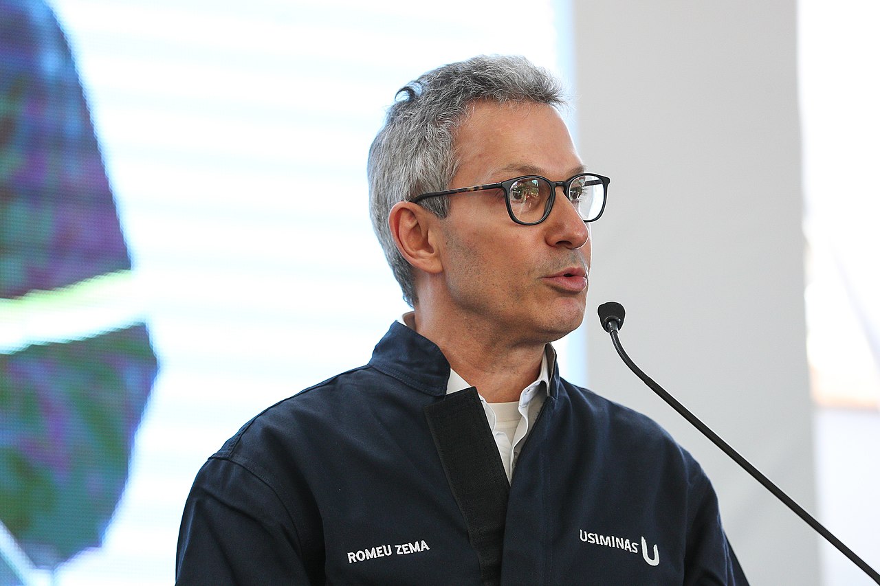 Governador de Minas Gerais, Romeu Zema (Novo) é um homem branco com cabelos grisalhos e olhos pretos. Na imagem ele usa óculos e um uniforme da Usiminas com seu nome escrito nele. Zema fala ao microfone e está virado de lado