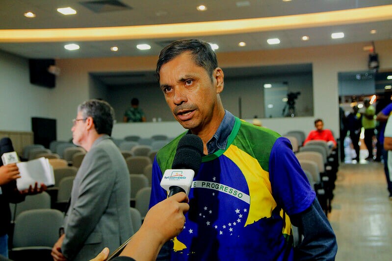Jailson Mesquita, coordenador político do MGL, é um homem negro de cabelos pretos e na imagem veste uma camiseta de mangas curtas com a estampa da bandeira do Brasil, ele dá uma entrevista.