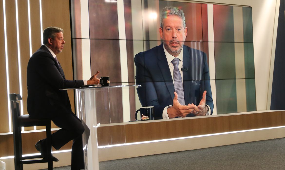 Presidente da Câmara, Arthur Lira, é um homem branco com cabelos e barbas grisalhos. Na imagem ele está sentado de lado e sua imagem é refletida em um telão atrás dele. Lira fala ao microfone e gesticula