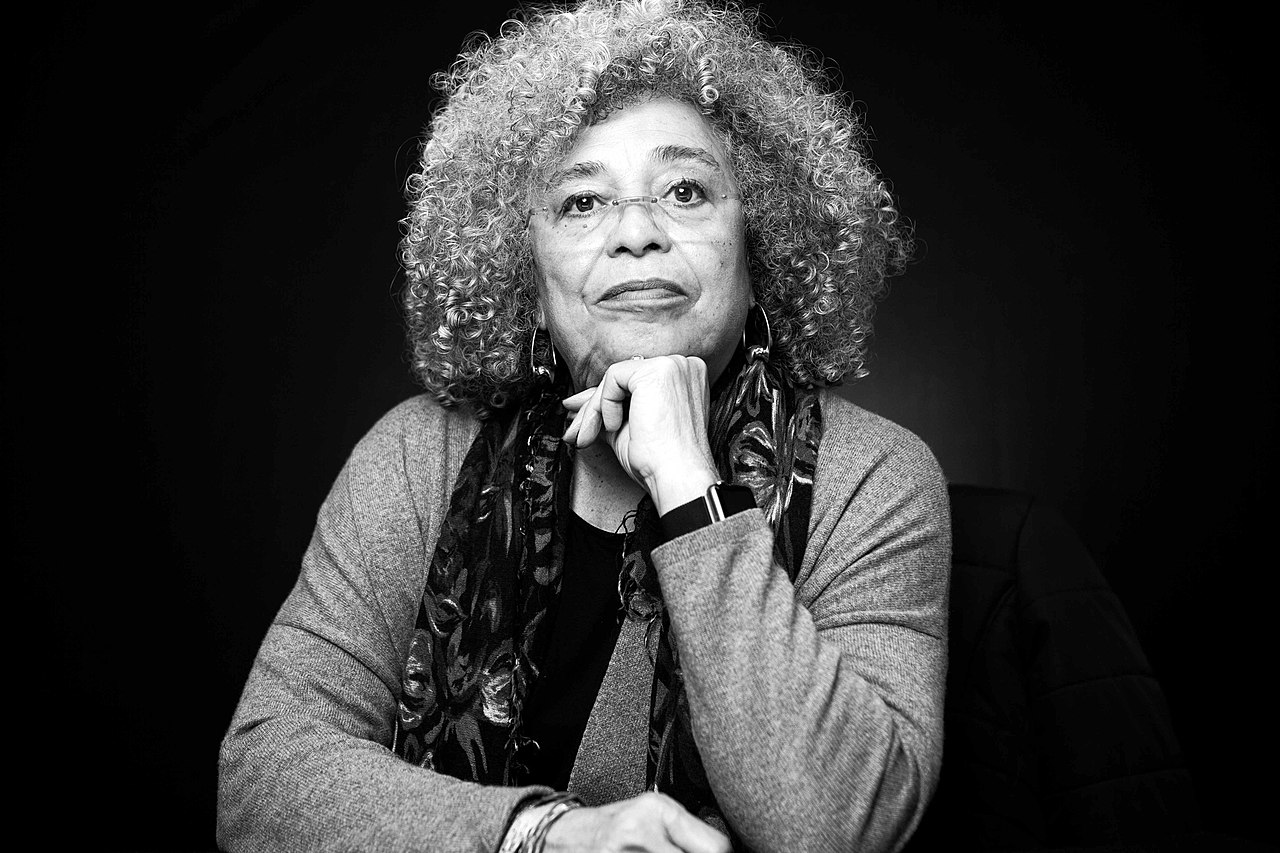 Angela Davis é uma mulher negra com cabelos cacheados, ela usa óculos cachecol e camisa de mangas compridas. Angela posa para a foto com a mão no queixo.