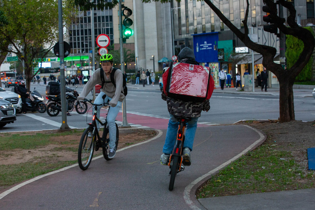 Na imagem entregadores de empresas de aplicativo andam em suas bicicletas a caminho de seus destinos.