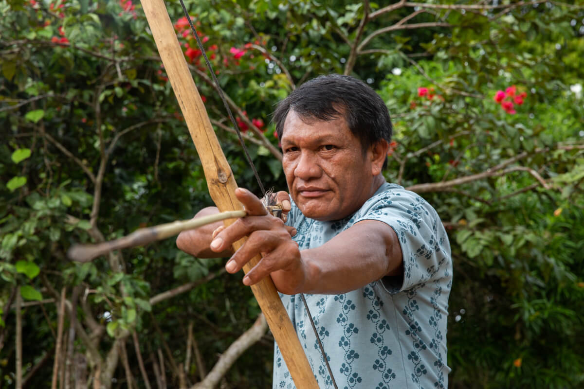 Na imagem, Olímpio Guajajara segura um arco e flecha. Ele veste uma camiseta cinza estampada.