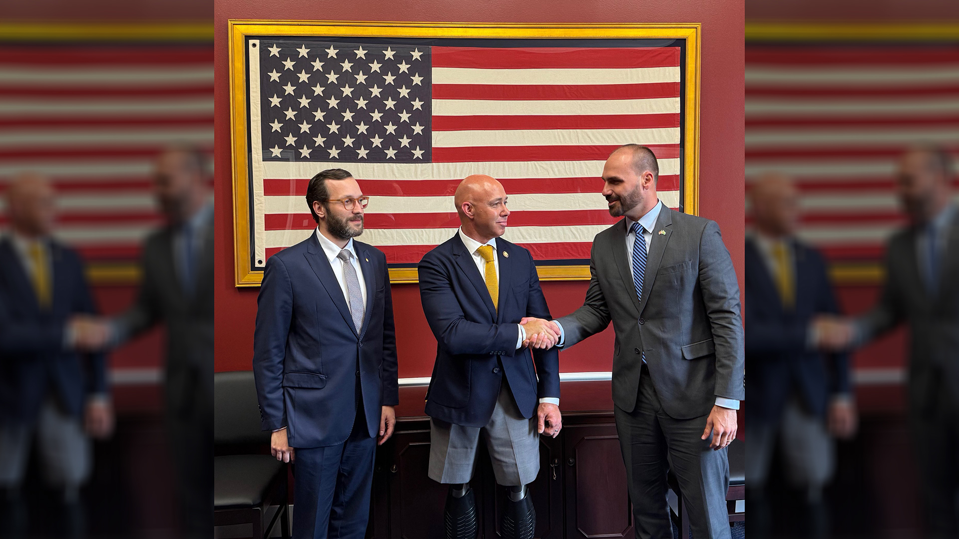 Eduardo Bolsonaro, ao centro-direita da imagem, aperta a mão de um parlamentar dos EUA durante encontro político; ao fundo, bandeira dos Estados Unidos emoldurada.