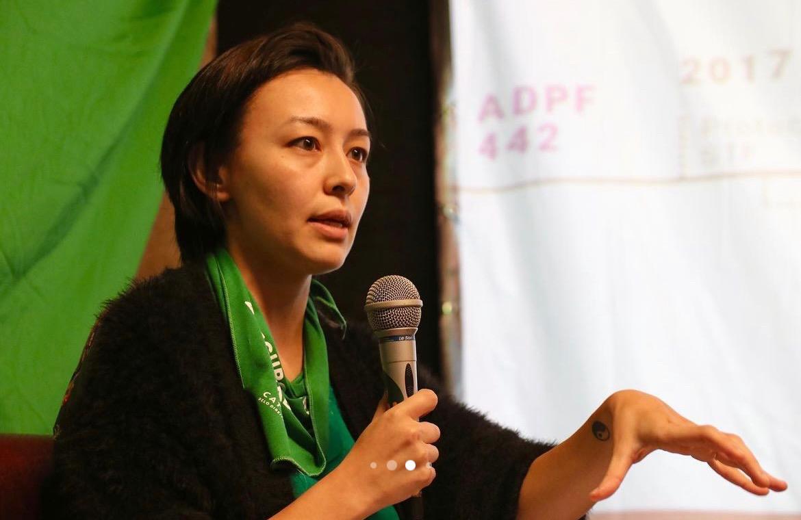 Advogada Letícia Ueda Vella com lenço verde no pescoço segura microfone enquanto fala em evento sobre direitos reprodutivos e descriminalização do aborto; ao fundo, faixa com referência à ADPF 442.
