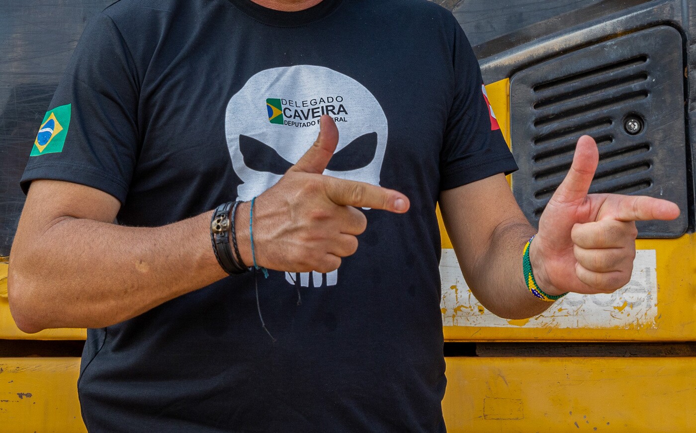 Homem usando camiseta preta com estampa de uma caveira e os dizeres “Delegado Caveira Deputado Federal”, fazendo gestos com as mãos diante de um veículo